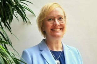 Halbporträt von Kristin Ruske; Destinationsleiterin Tourist-Information Lutherstadt Wittenberg im hellblauen Blazer, lächelnd mit blonden kurzen Haaren und Brille