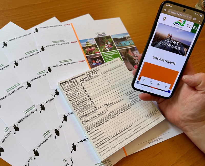 Handy, das die digitale Gästekarte auf demm Handy zeigt, alte Papiergästekarten ligen im Hintergrund