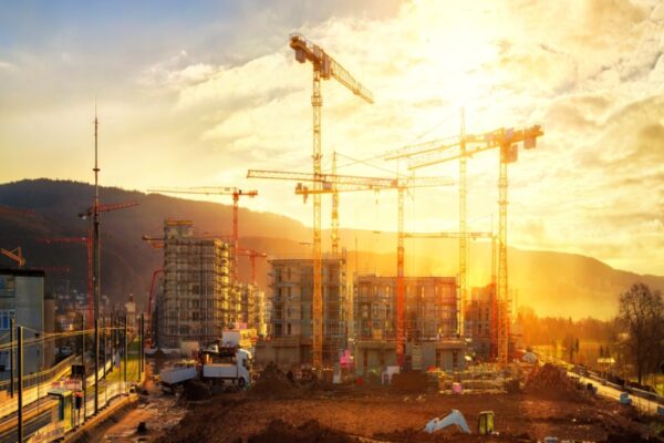 Große Baustelle mit mehreren Kränen, die an einem Gebäudekomplex arbeiten, beleuchtet von warmem, goldenem Sonnenlicht.