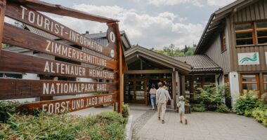Informationstafel der Tourist-Information Altenau aus Holz links im Bild und Famile Mann, Frau & zwei KInder, die zum Eingang des Holzhauses der Tourist-Information Altenau laufen